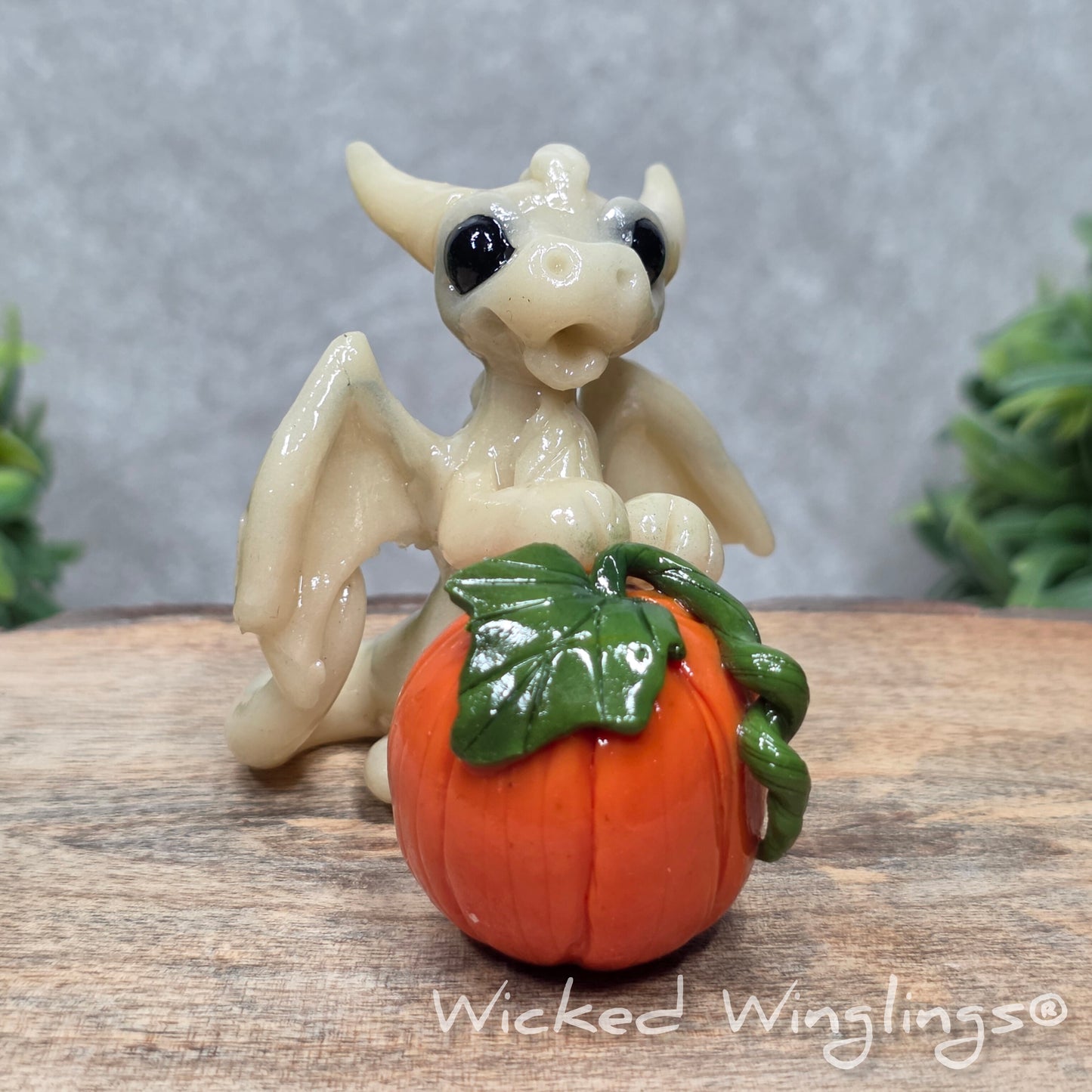 A baby clay dragon sculpture with a pumpkin against a wood background with greenery and text Wicked Winglings