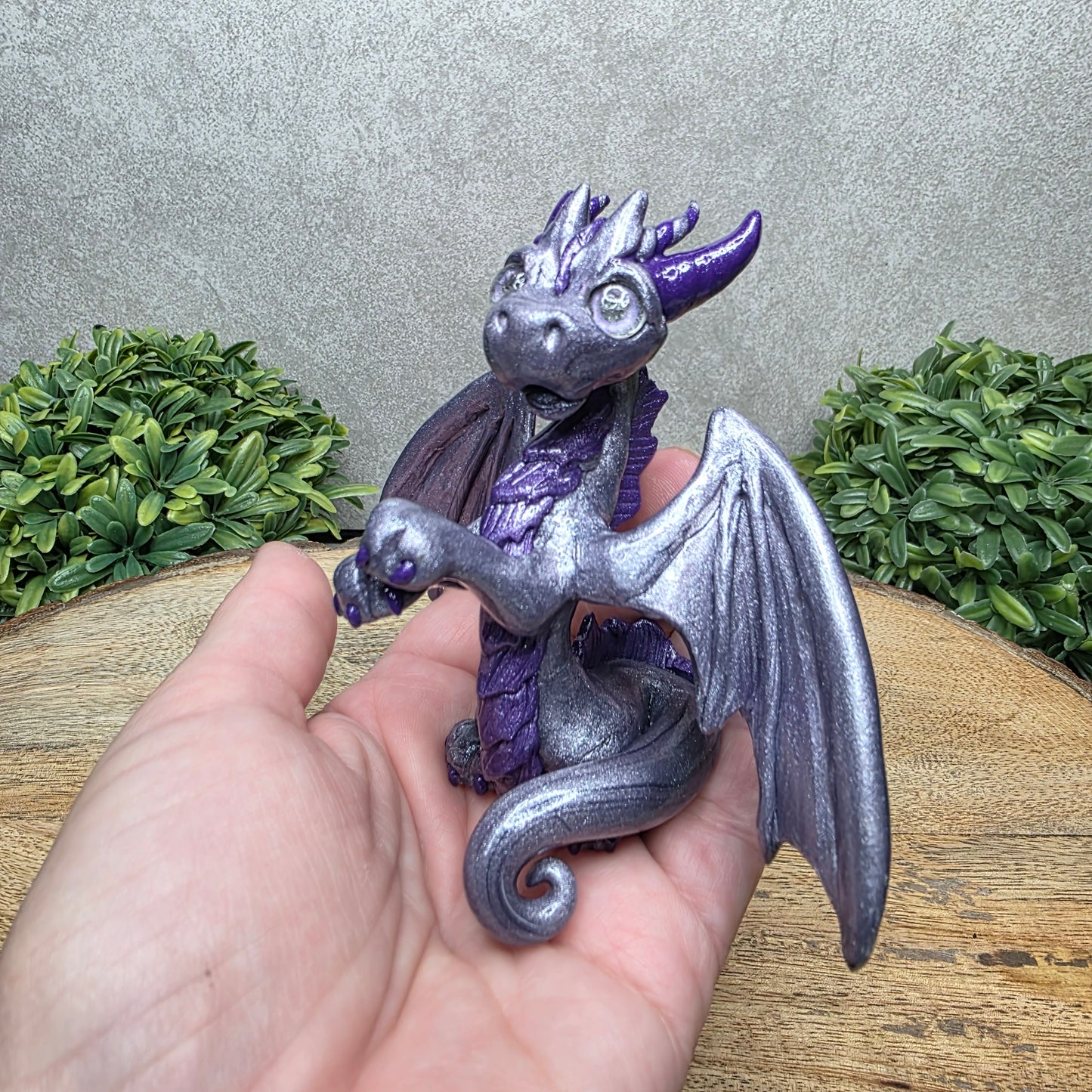 A hand holding a clay dragon against a wood background with greenery