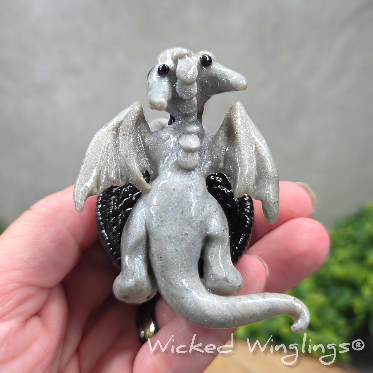 A hand holding a baby clay dragon with a cookie with a Wicked Winglings® watermark.