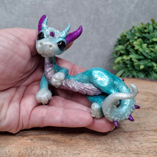A hand holding a clay dragon against a wood background with greenery