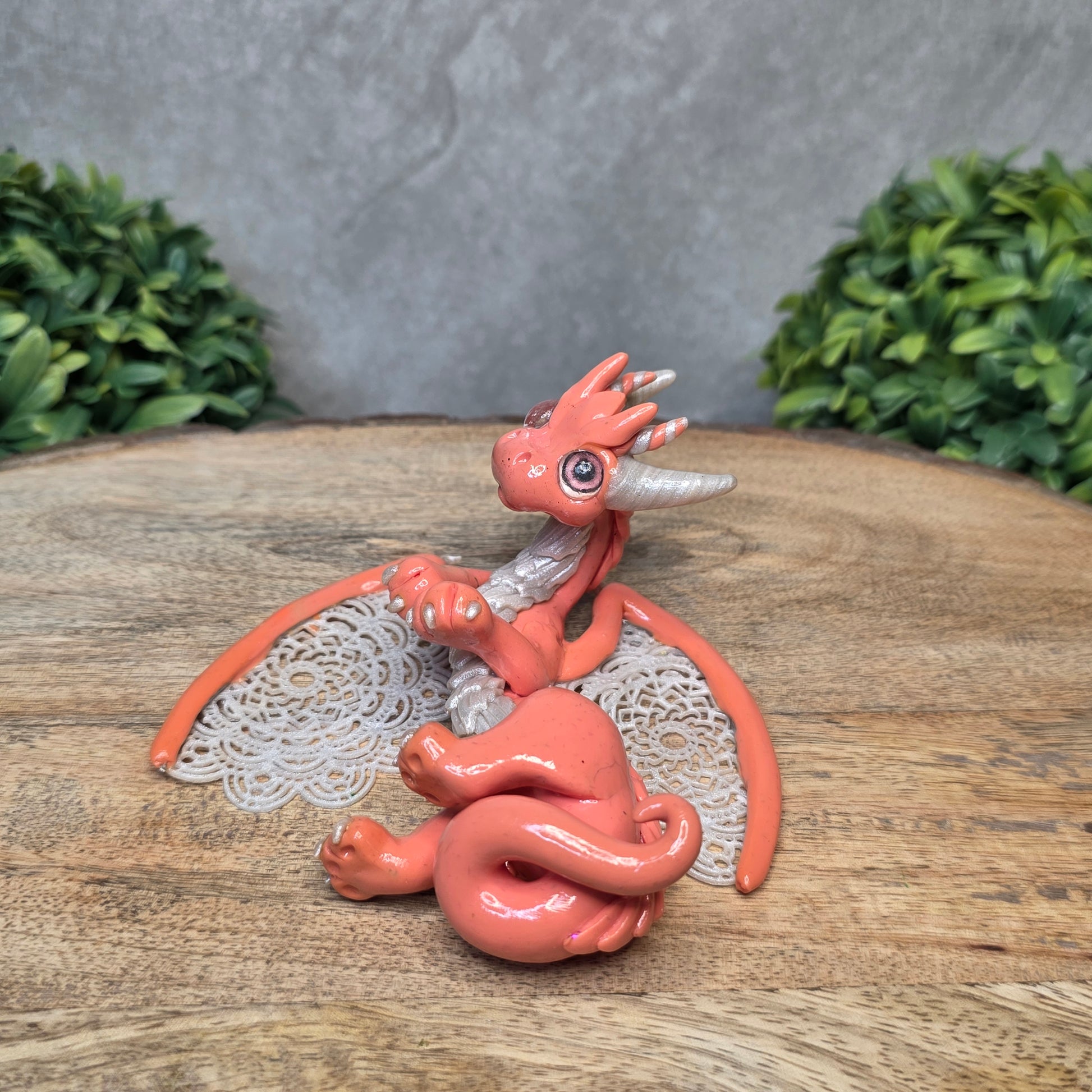 A clay dragon sculpture with lacy wings on a wood surface with greenery.