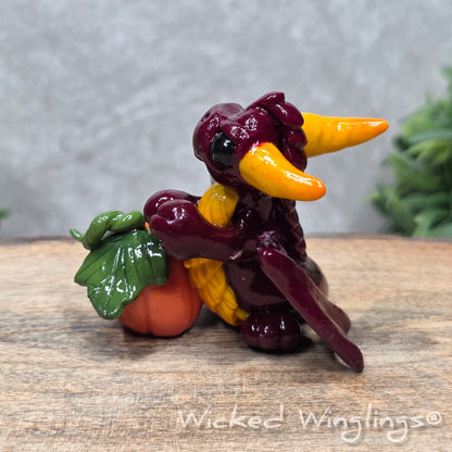 A baby clay dragon sculpture with a pumpkin against a wood background with greenery and text Wicked Winglings
