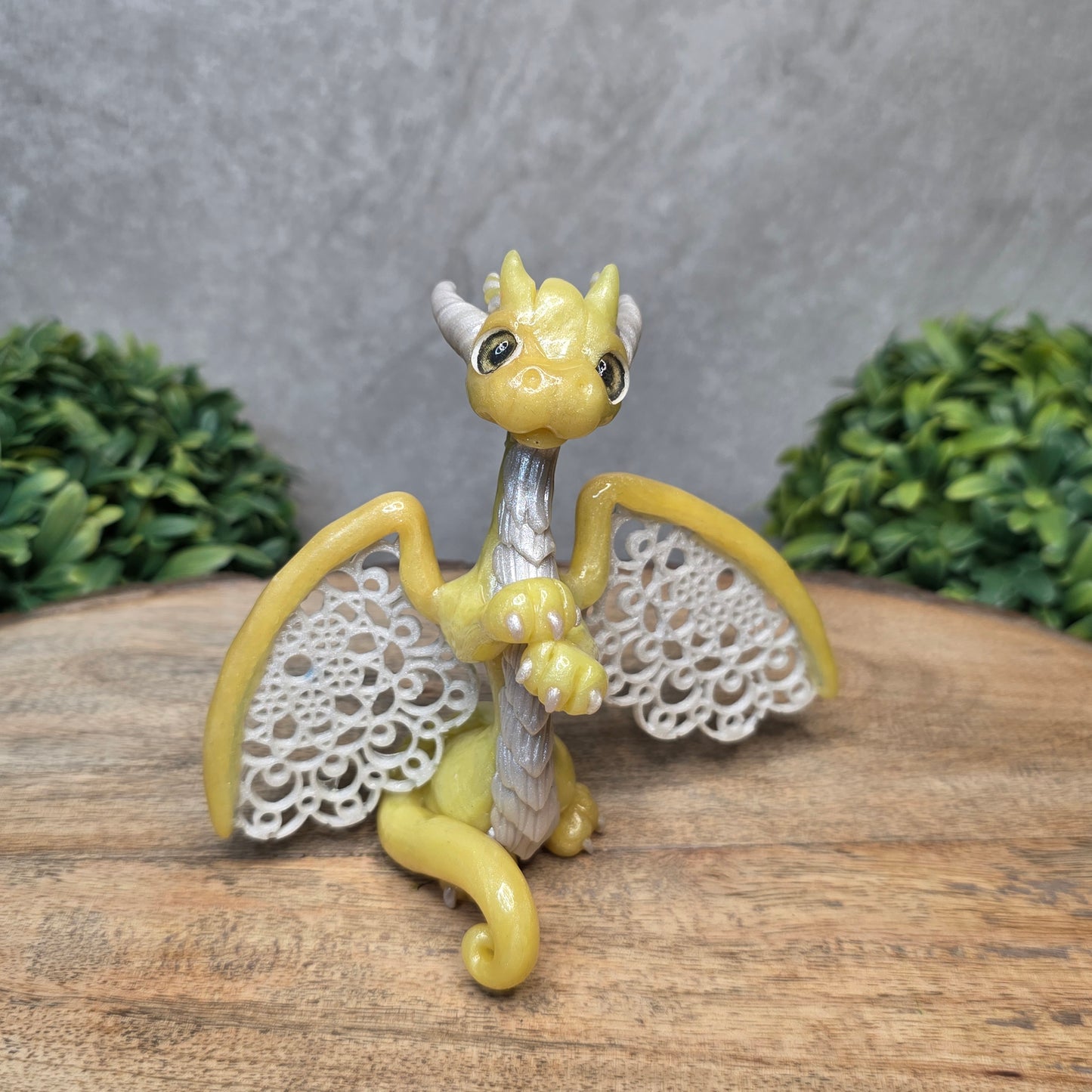 A clay dragon sculpture with lacy wings on a wood surface with greenery.