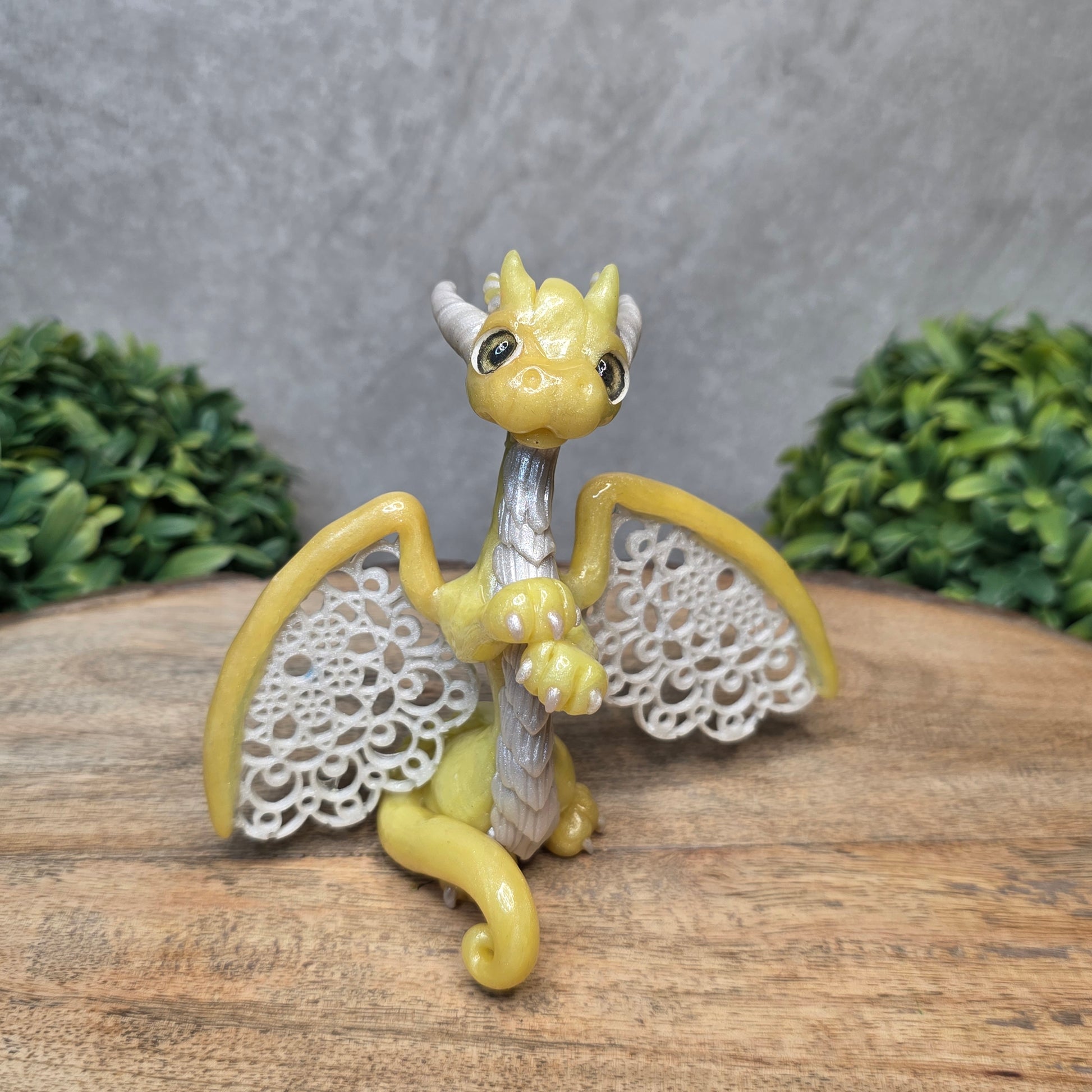 A clay dragon sculpture with lacy wings on a wood surface with greenery.