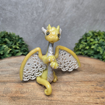 A clay dragon sculpture with lacy wings on a wood surface with greenery.