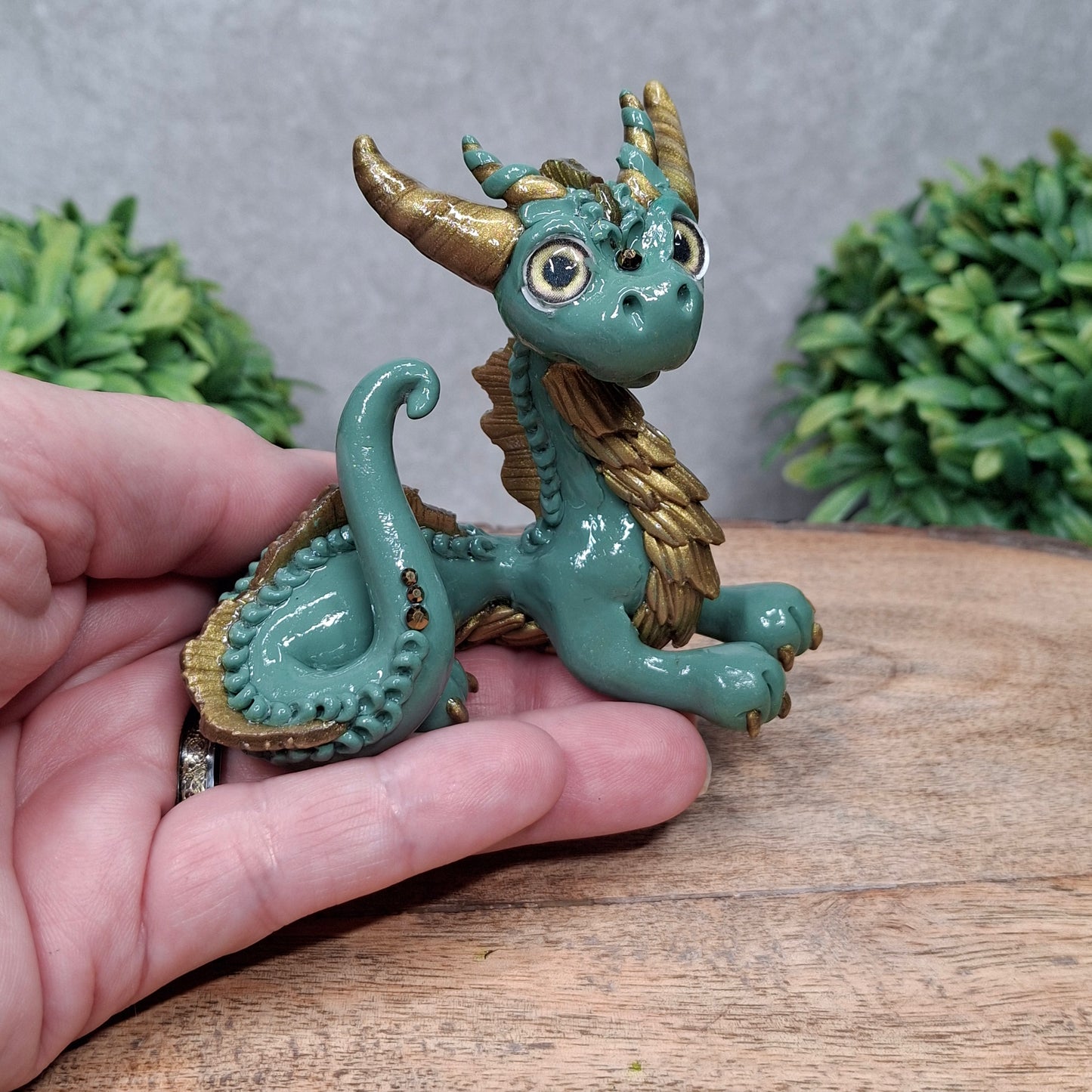 A hand holding a clay dragon against a wood background with greenery