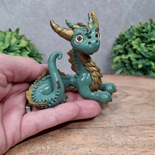 A hand holding a clay dragon against a wood background with greenery