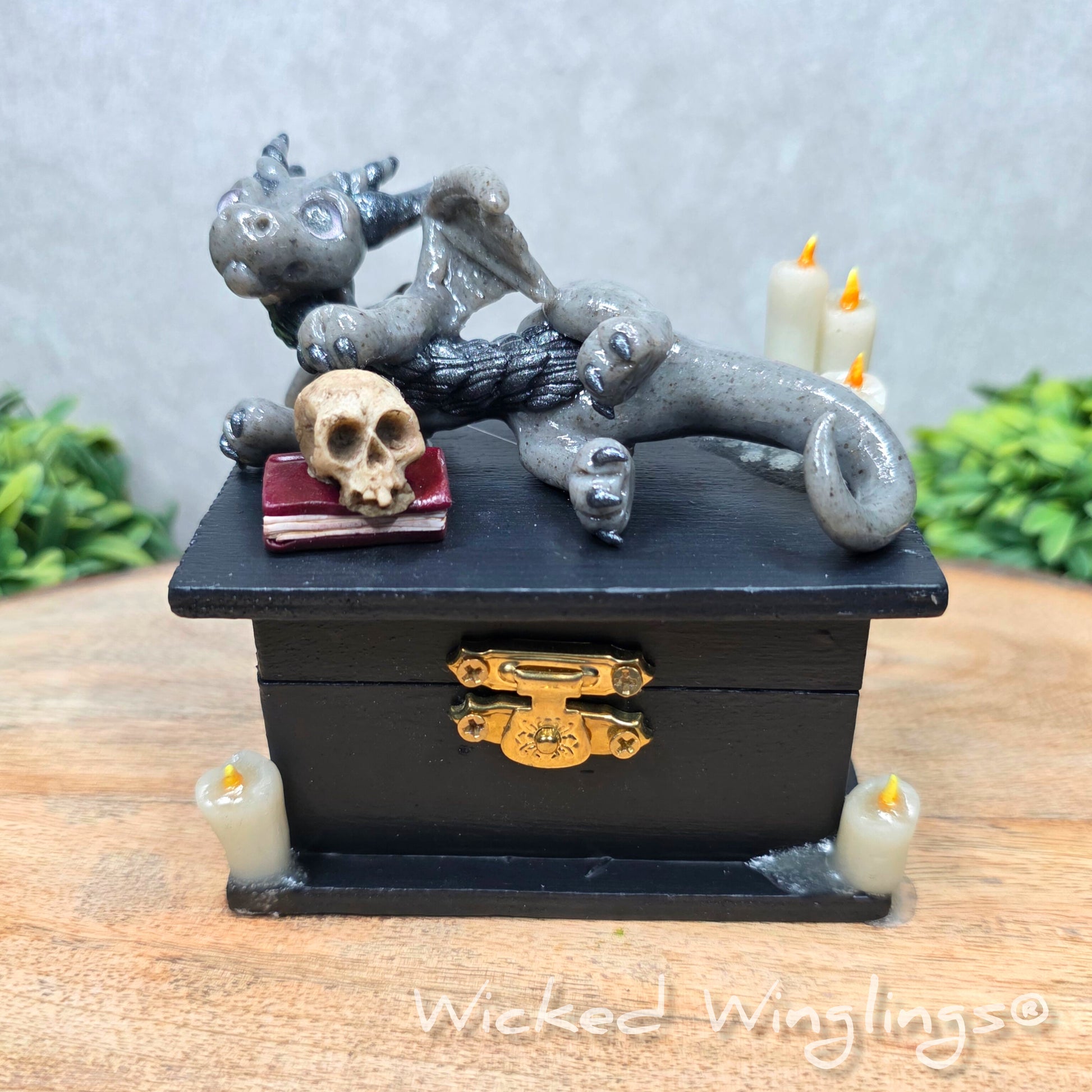 A clay dragon on a black box with candles and a skull on a wood surface with greenery. Includes text Wicked Winglings