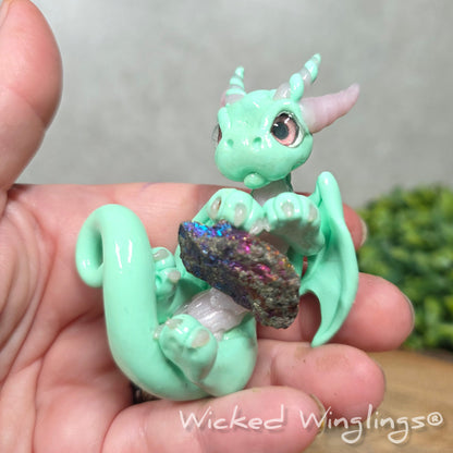 A hand holding a mint clay dragon holding a rainbow colored stone against a blurred background with greenery. Text says Wicked Winglinng.