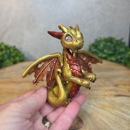 A hand holding a gold and bronze clay dragon against a blurred wood surface