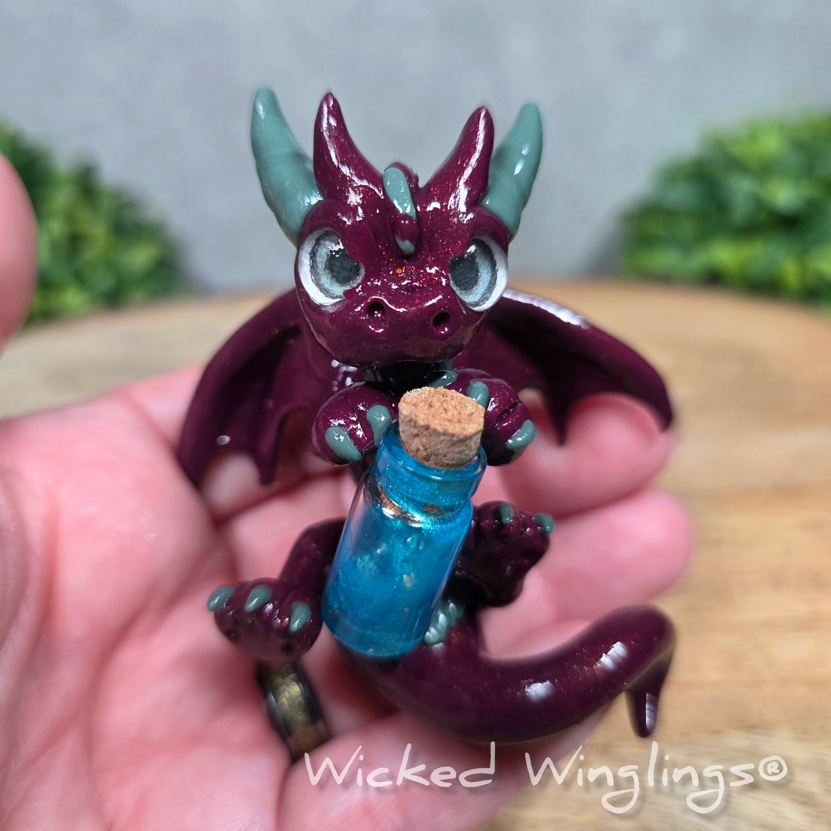 A hand holding a clay dragon with a potion bottle. Text reads Wicked Winglings.