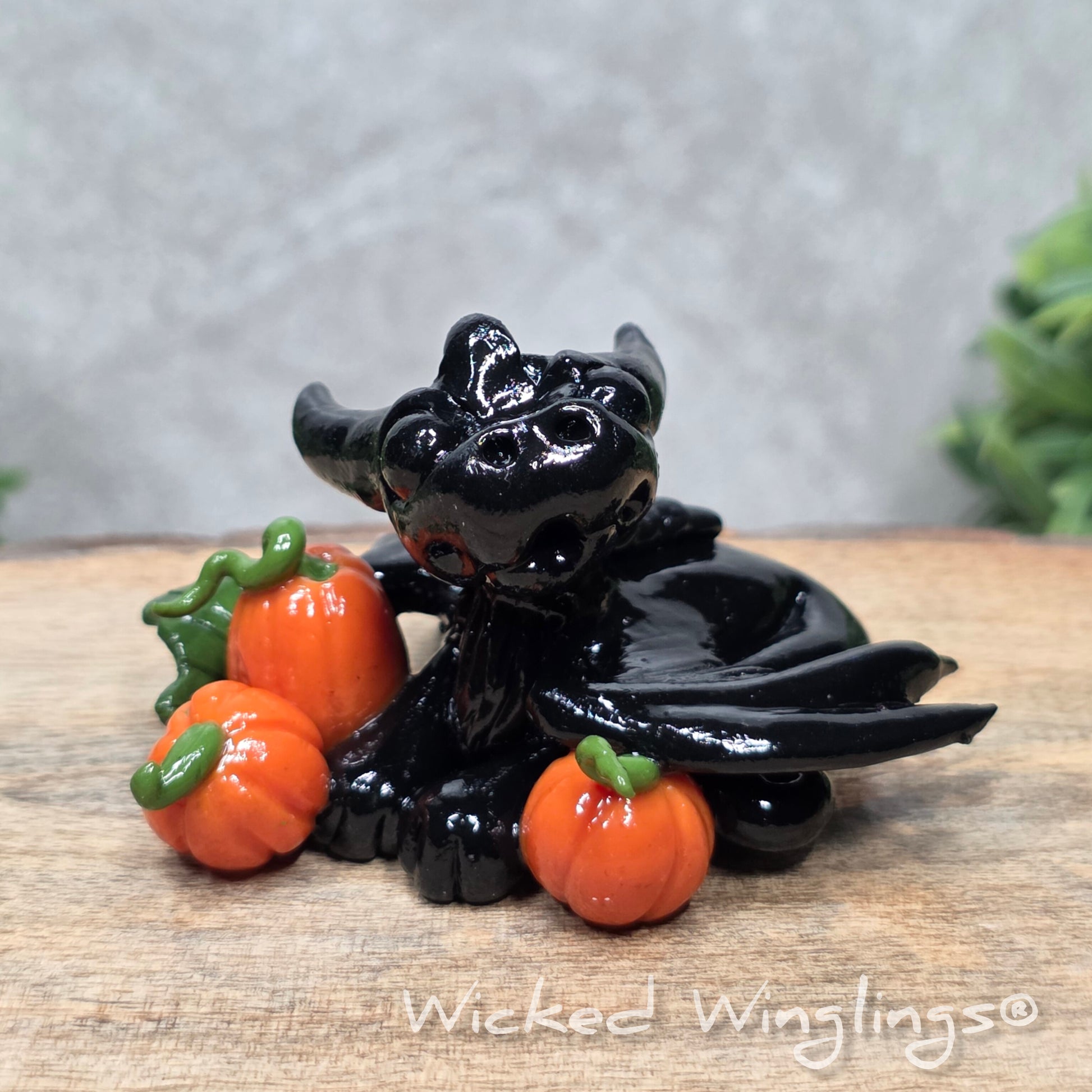 A baby clay dragon sculpture with pumpkins against a wood background with greenery and text Wicked Winglings