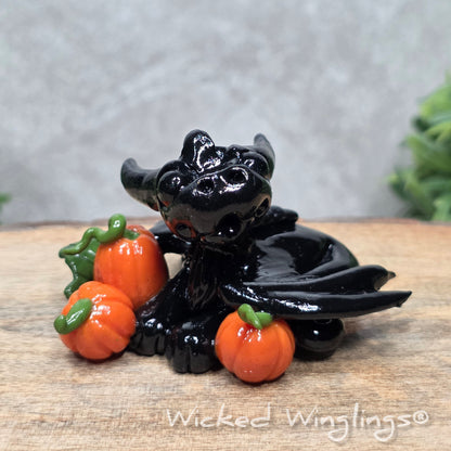 A baby clay dragon sculpture with pumpkins against a wood background with greenery and text Wicked Winglings