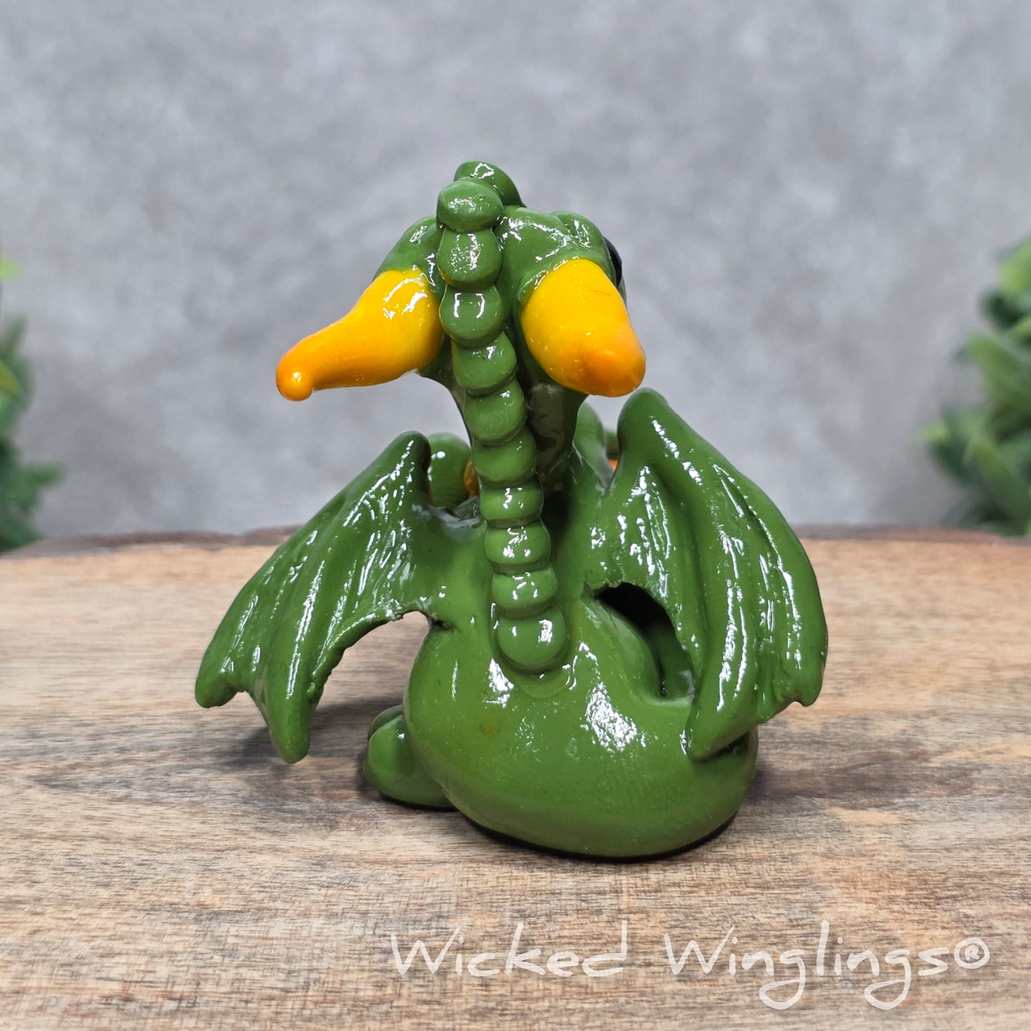 A baby clay dragon sculpture with pumpkins against a wood background with greenery and text Wicked Winglings