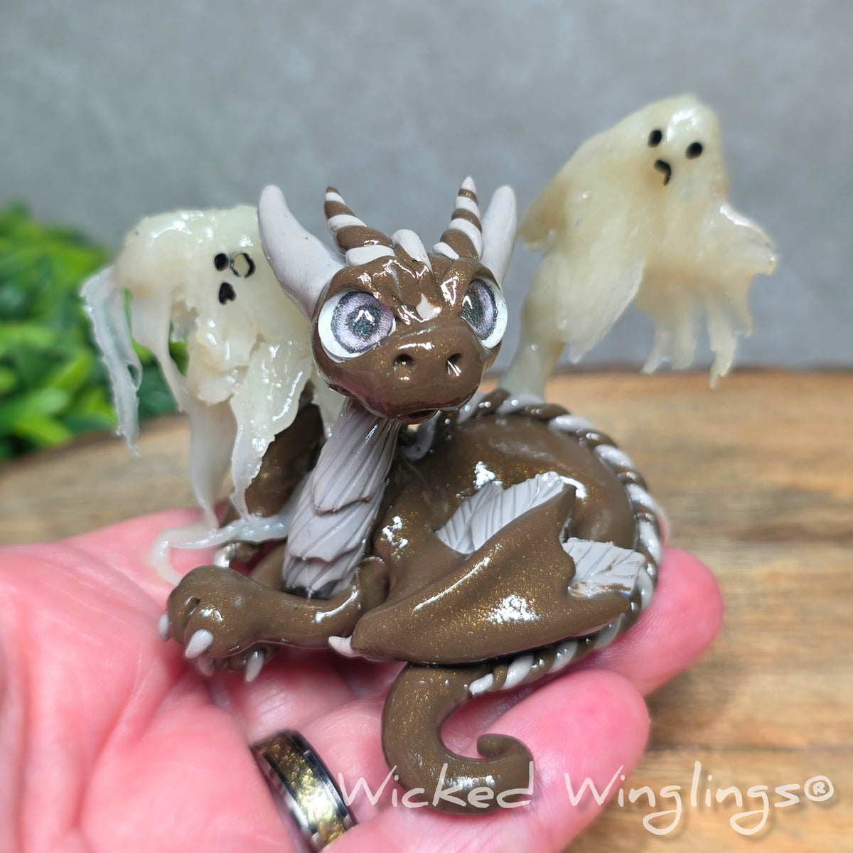 A hand holding a brown clay dragon with ghosts against a wood surface with greenery. Text reads Wicked Winglings.