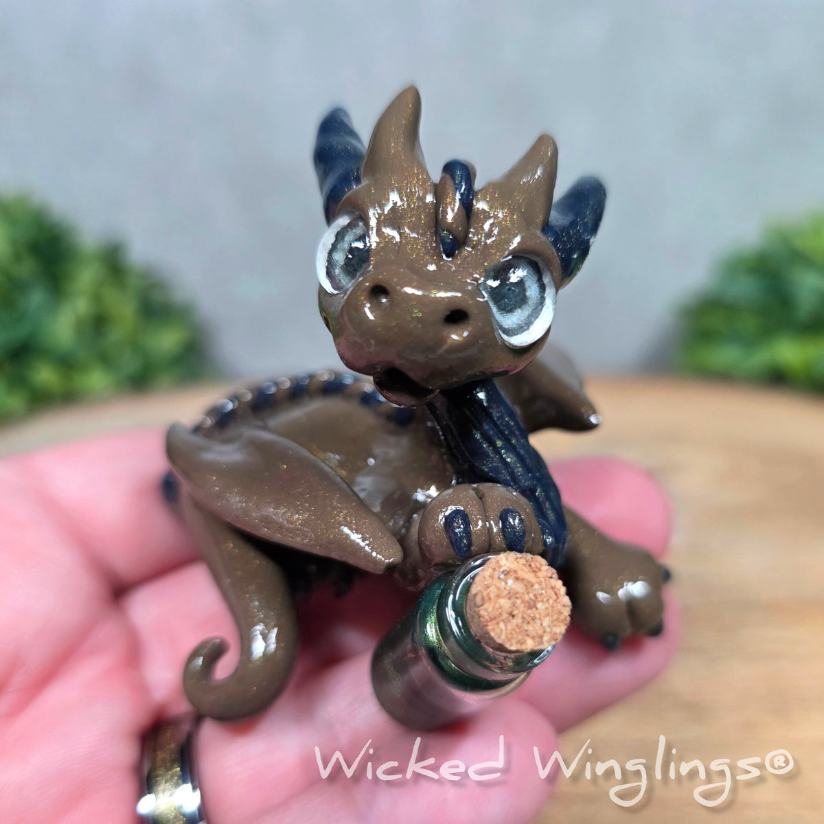A hand holding a clay dragon with a potion bottle on a wood surface. Text reads Wicked Winglings.