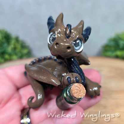 A hand holding a clay dragon with a potion bottle on a wood surface. Text reads Wicked Winglings.