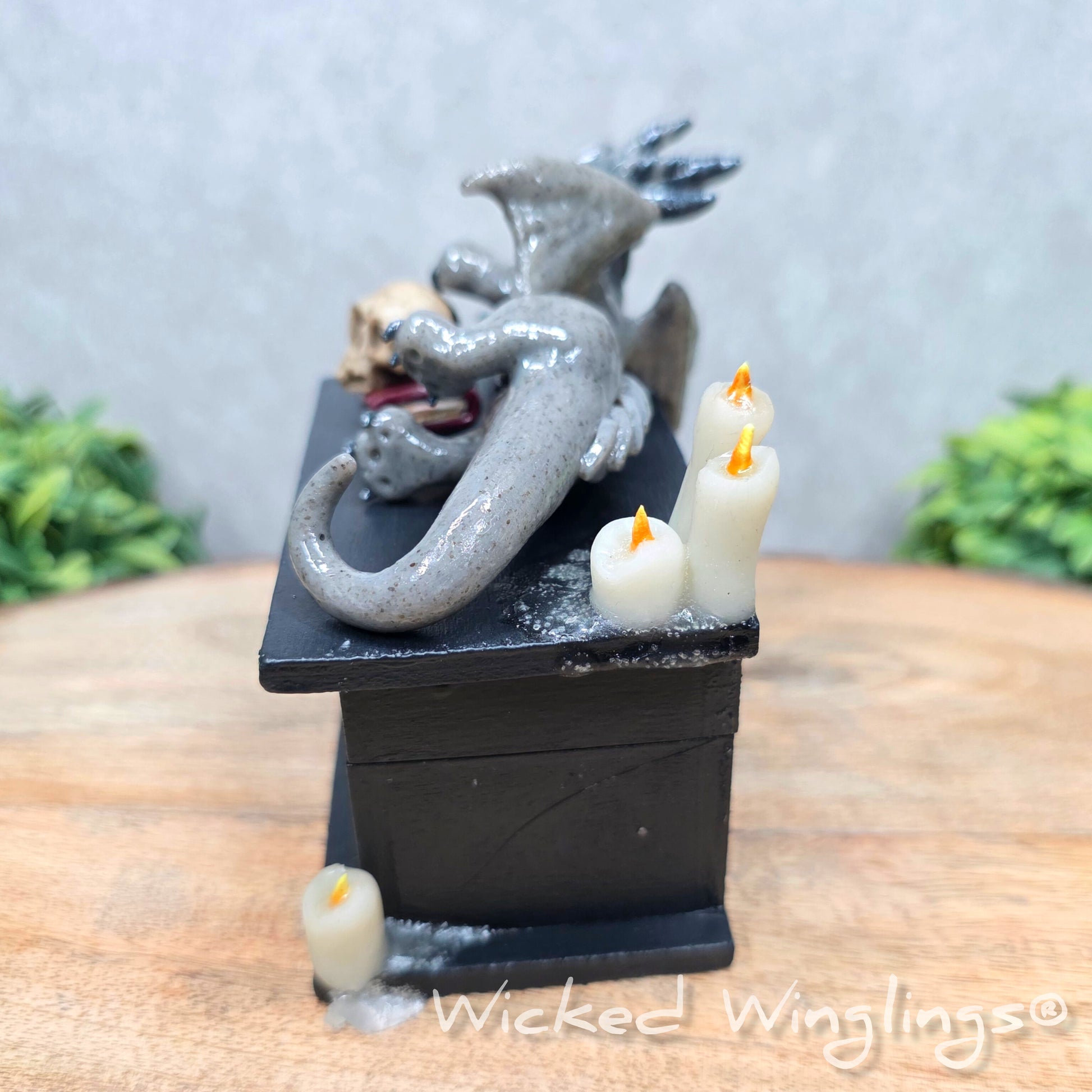A clay dragon on a black box with candles and a skull on a wood surface with greenery. Includes text Wicked Winglings