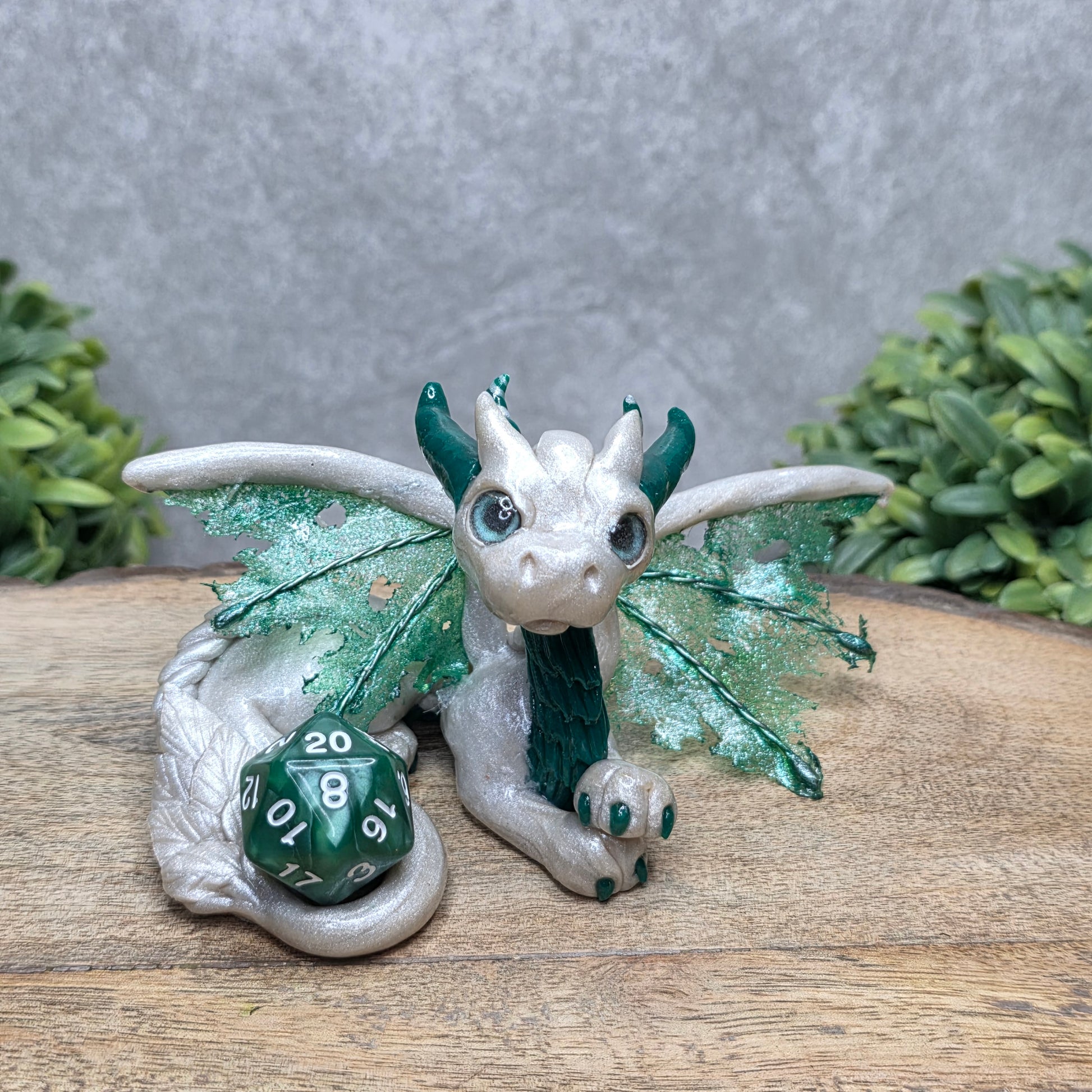 Aclay dragon with a D20 dice against a wood surface with greenery.