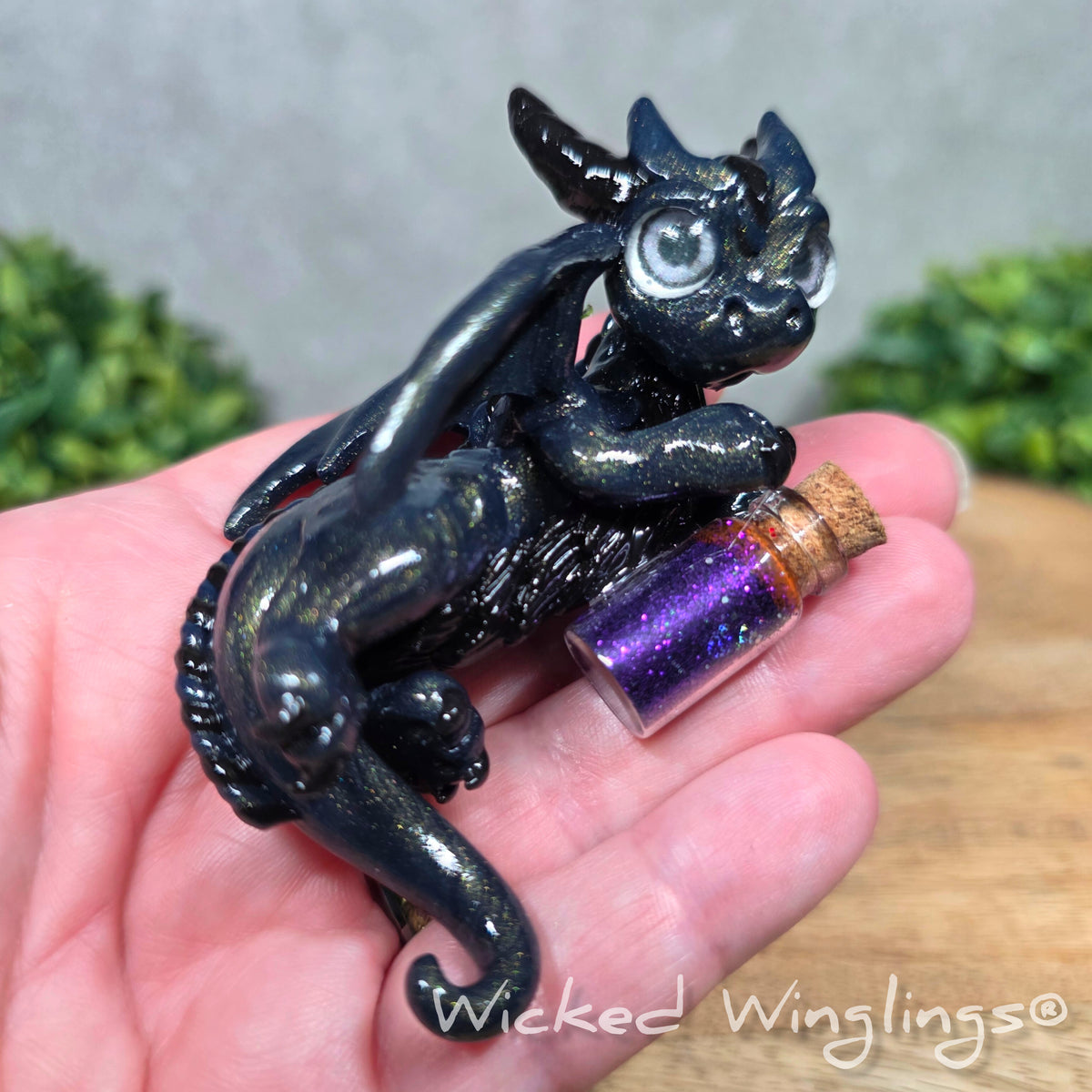 A hand holding a clay dragon with a potion bottle on a wood surface. Text reads Wicked Winglings.