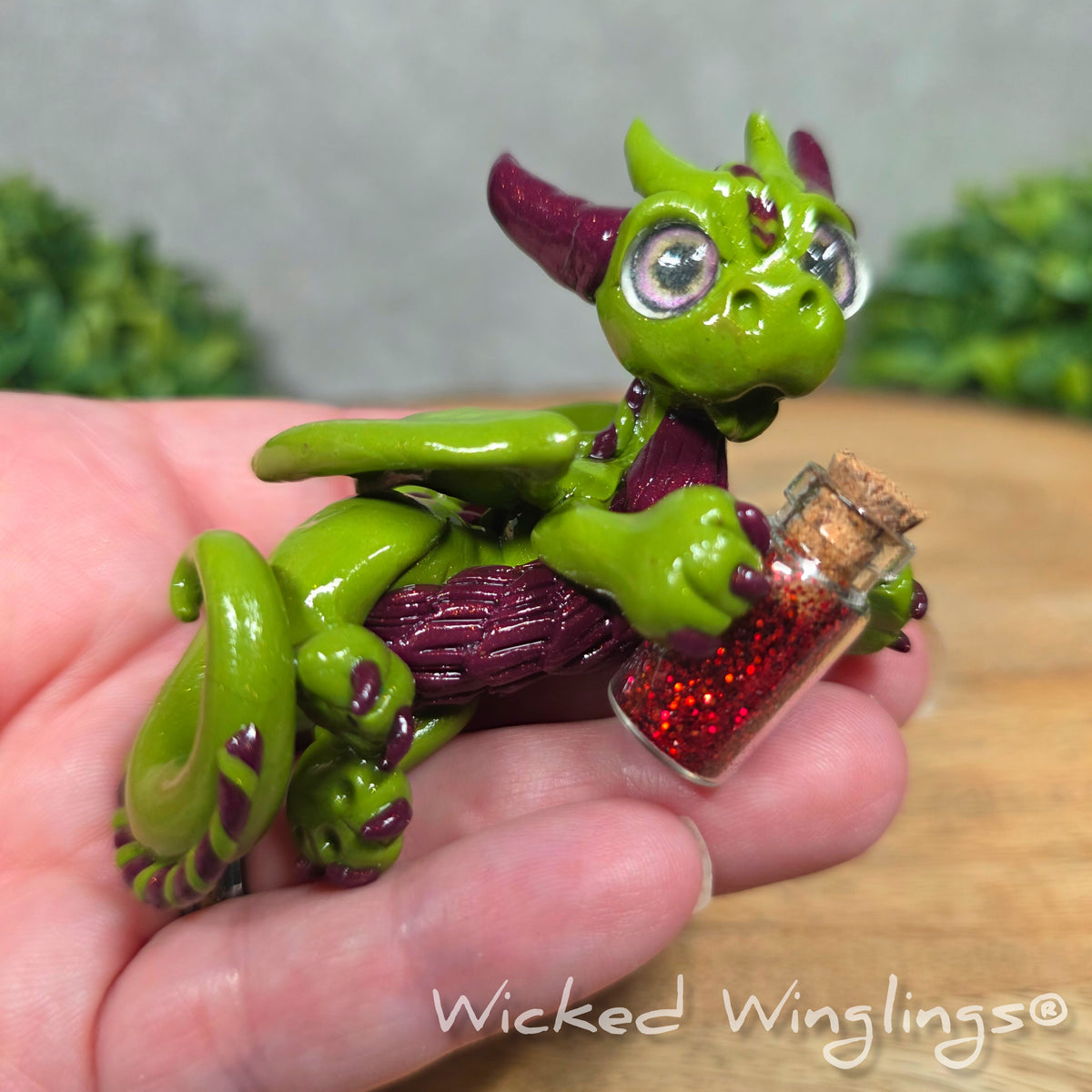 A hand holding a clay dragon with a potion bottle on a wood surface. Text reads Wicked Winglings.