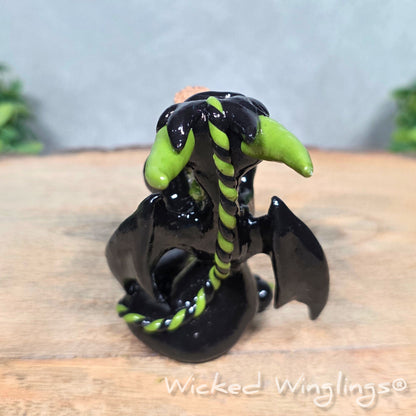A clay dragon with a potion bottle on a wood surface. Text reads Wicked Winglings.