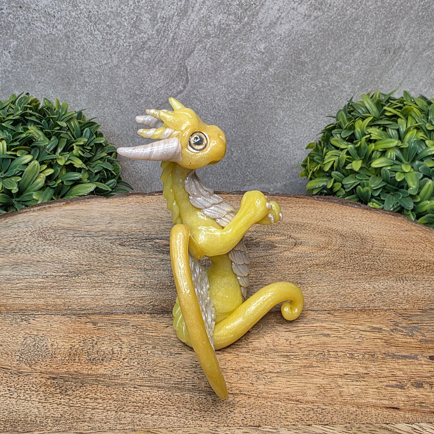 A clay dragon sculpture with lacy wings on a wood surface with greenery.