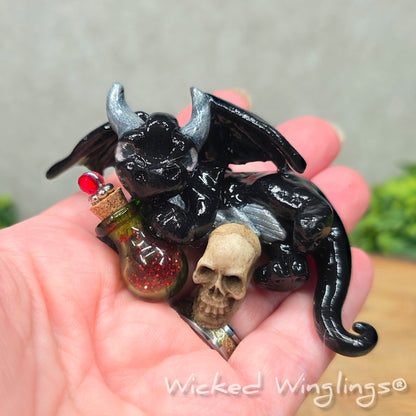 A hand holding a clay dragon with a skull and potion bottle on a wood surface with greenery. Includes text Wicked Winglings