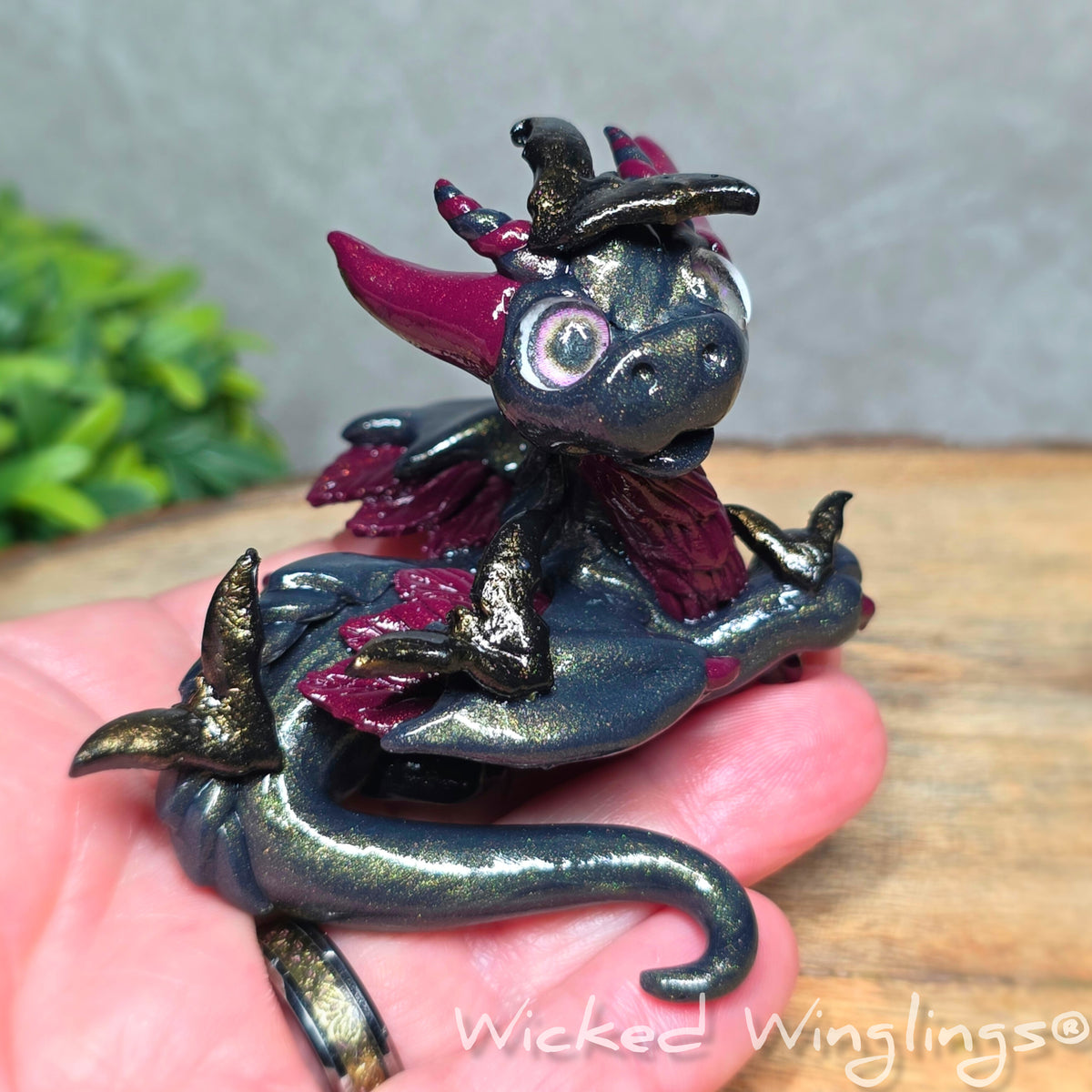 A hand holding a clay dragon with bats against a wood surface with greenery. Text reads Wicked Winglings.