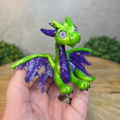 A hand holding a green and purple clay dragon against a blurred wood surface