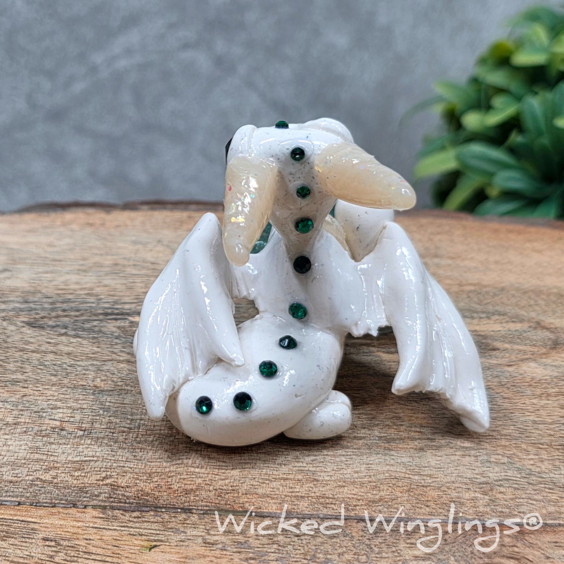 A baby clay dragon sculpture with a crackled quartz against a wood background with greenery and text Wicked Winglings
