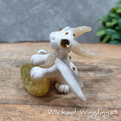 A baby clay dragon sculpture with a crackled quartz against a wood background with greenery and text Wicked Winglings