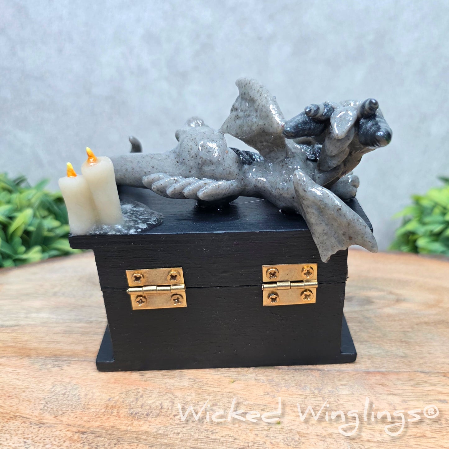 A clay dragon on a black box with candles and a skull on a wood surface with greenery. Includes text Wicked Winglings