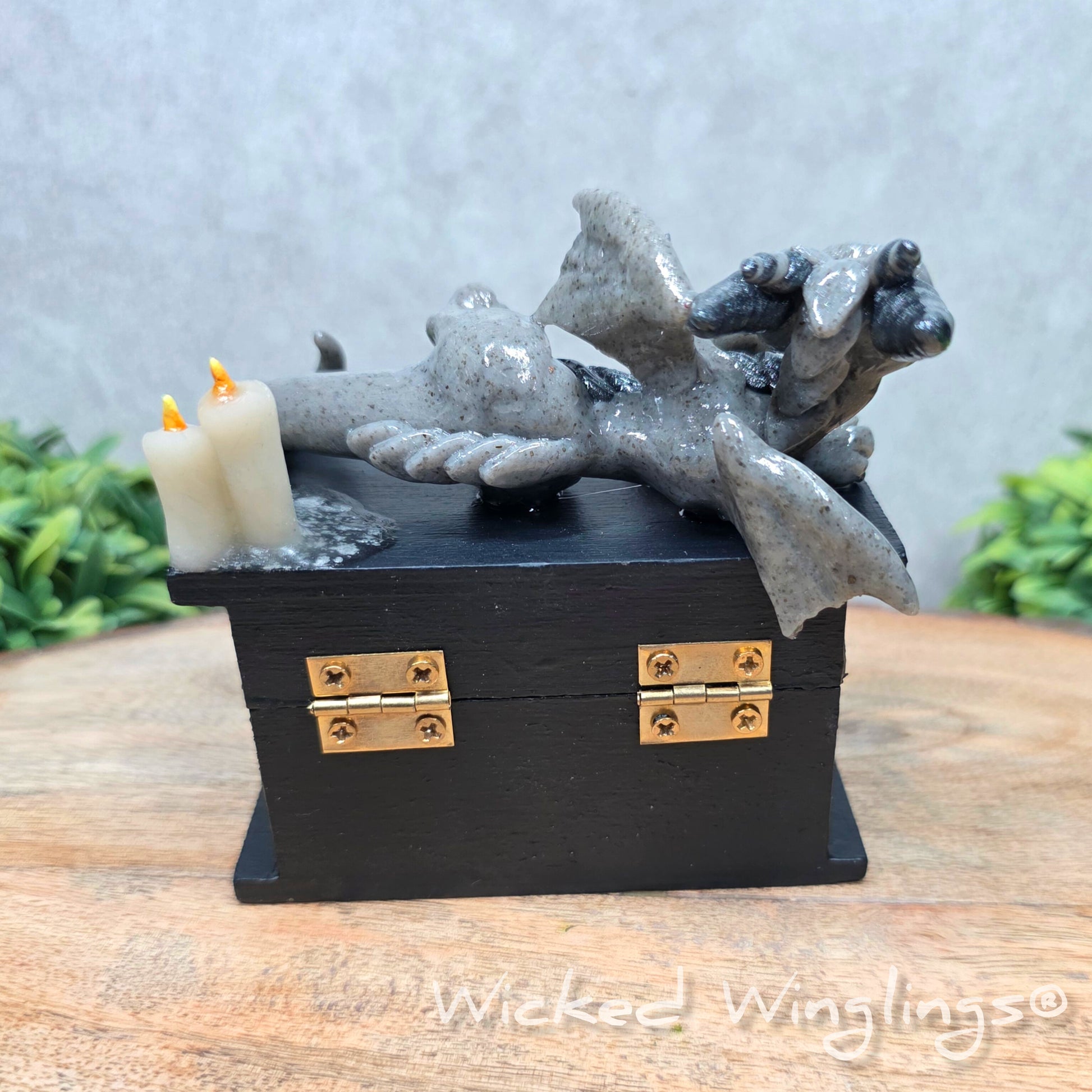 A clay dragon on a black box with candles and a skull on a wood surface with greenery. Includes text Wicked Winglings