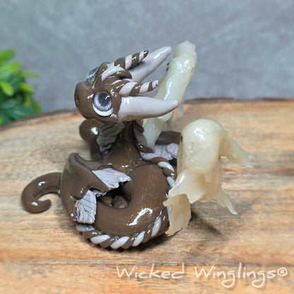 Side view of a  brown clay dragon with ghosts on a wood surface with greenery. Text reads Wicked Winglings.