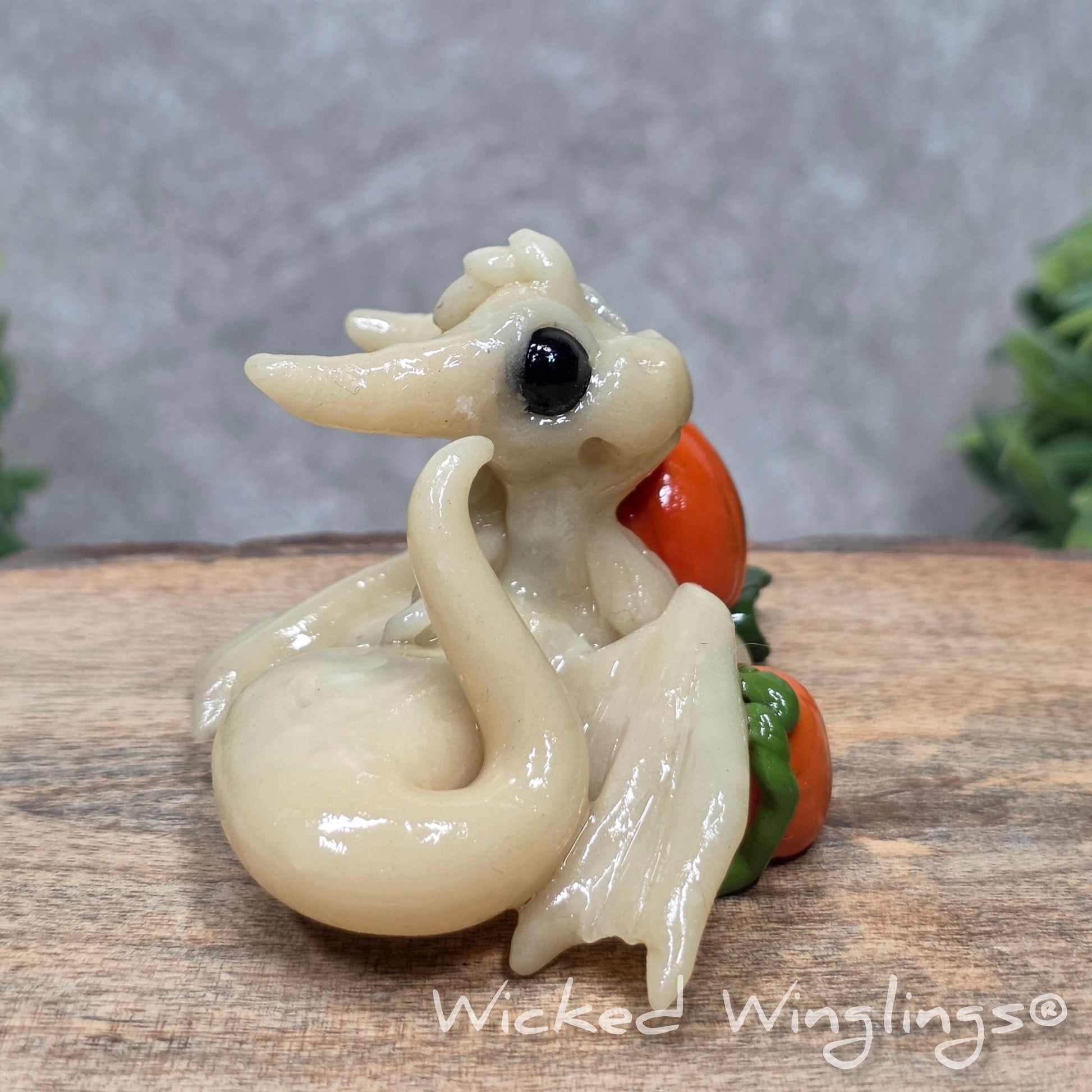 A baby clay dragon sculpture with pumpkins against a wood background with greenery and text Wicked Winglings