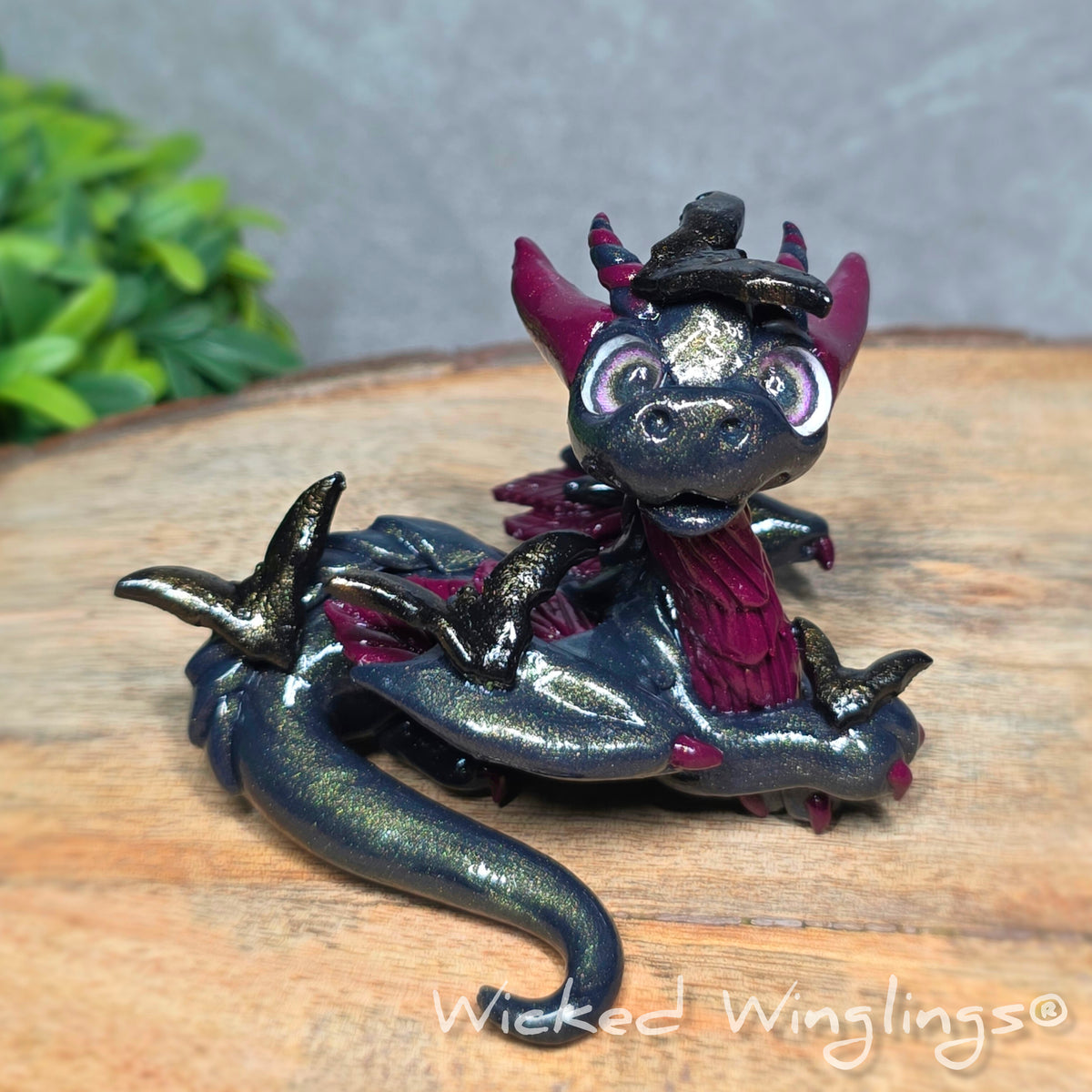 A clay dragon with bats sitting on a wood surface with greenery. Text reads Wicked Winglings.