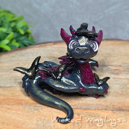 A clay dragon with bats sitting on a wood surface with greenery. Text reads Wicked Winglings.