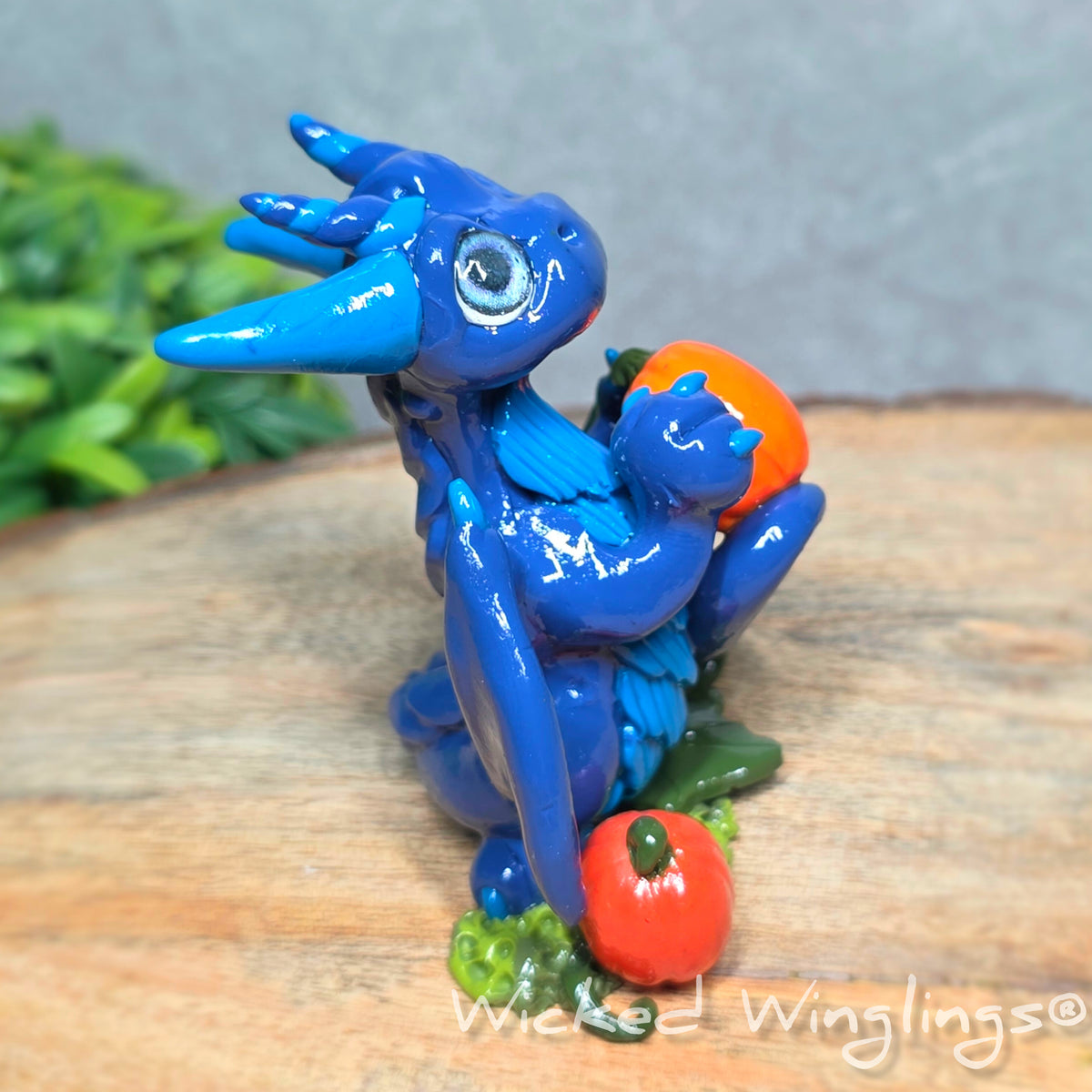 Side view of a blue clay dragon with pumpkins against a wood surface with greenery. Text reads Wicked Winglings. 
