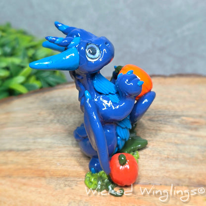 Side view of a blue clay dragon with pumpkins against a wood surface with greenery. Text reads Wicked Winglings. 