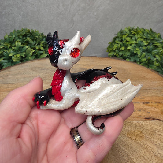 A hand holding a 2 toned clay dragon sculpture on a wood surface with greenery.