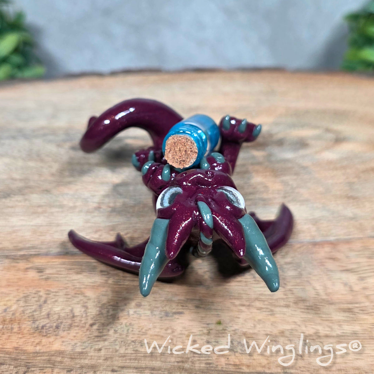 A clay dragon with a potion bottle on a wood surface. Text reads Wicked Winglings.