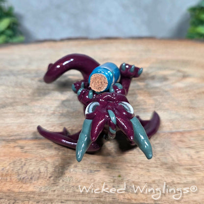 A clay dragon with a potion bottle on a wood surface. Text reads Wicked Winglings.