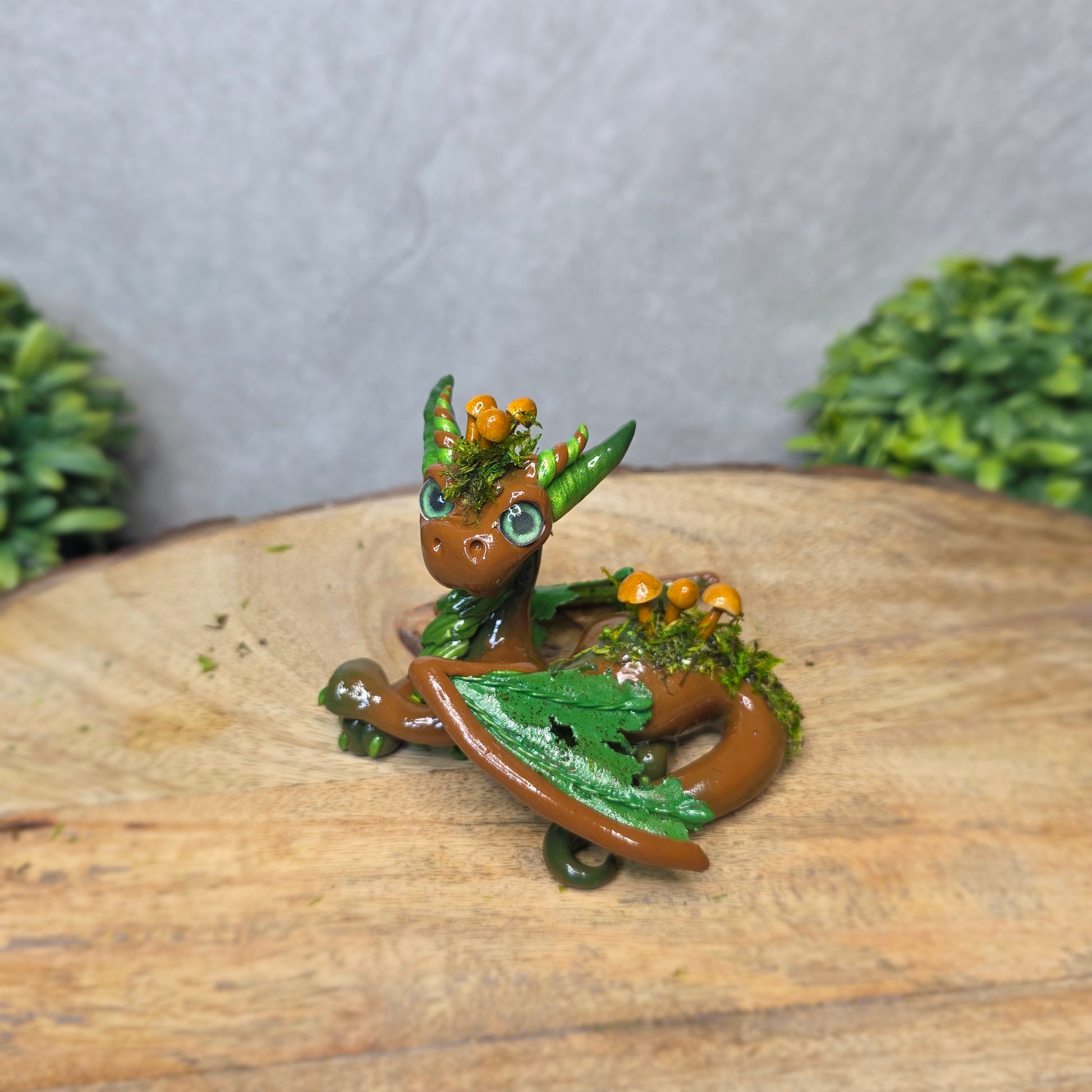 A clay dragon accented with moss and mushrooms against a wood surface with greenery.
