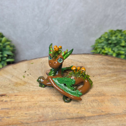 A clay dragon accented with moss and mushrooms against a wood surface with greenery.