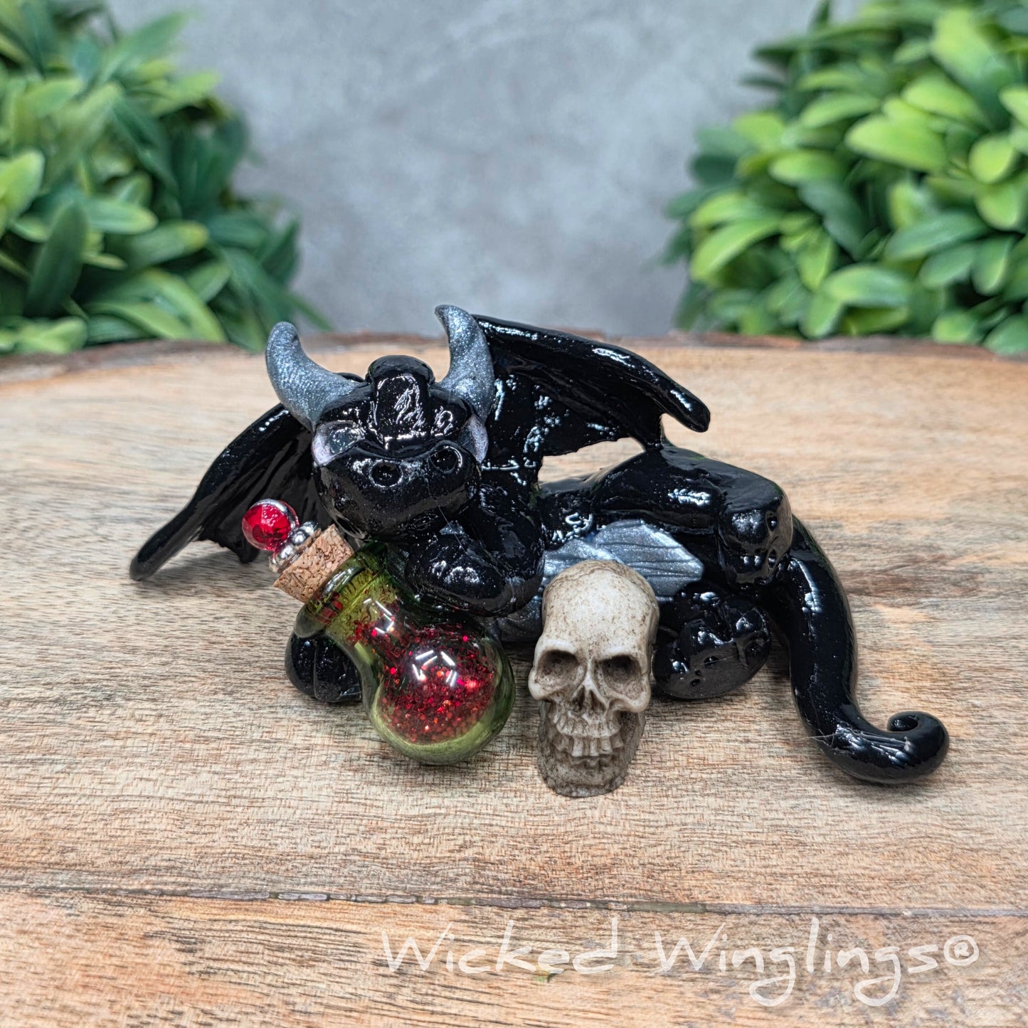 A clay dragon with a skull and potion bottle on a wood surface with greenery. Includes text Wicked Winglings