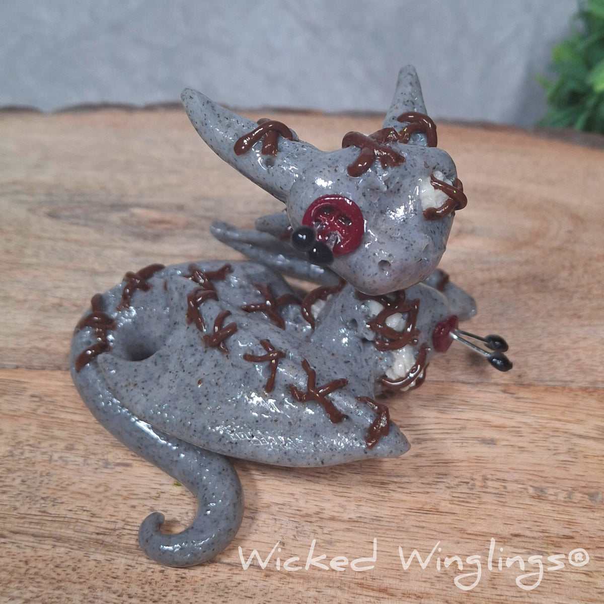A clay voodoo dragon on a wood surface with greenery. Text says Wicked Winglings