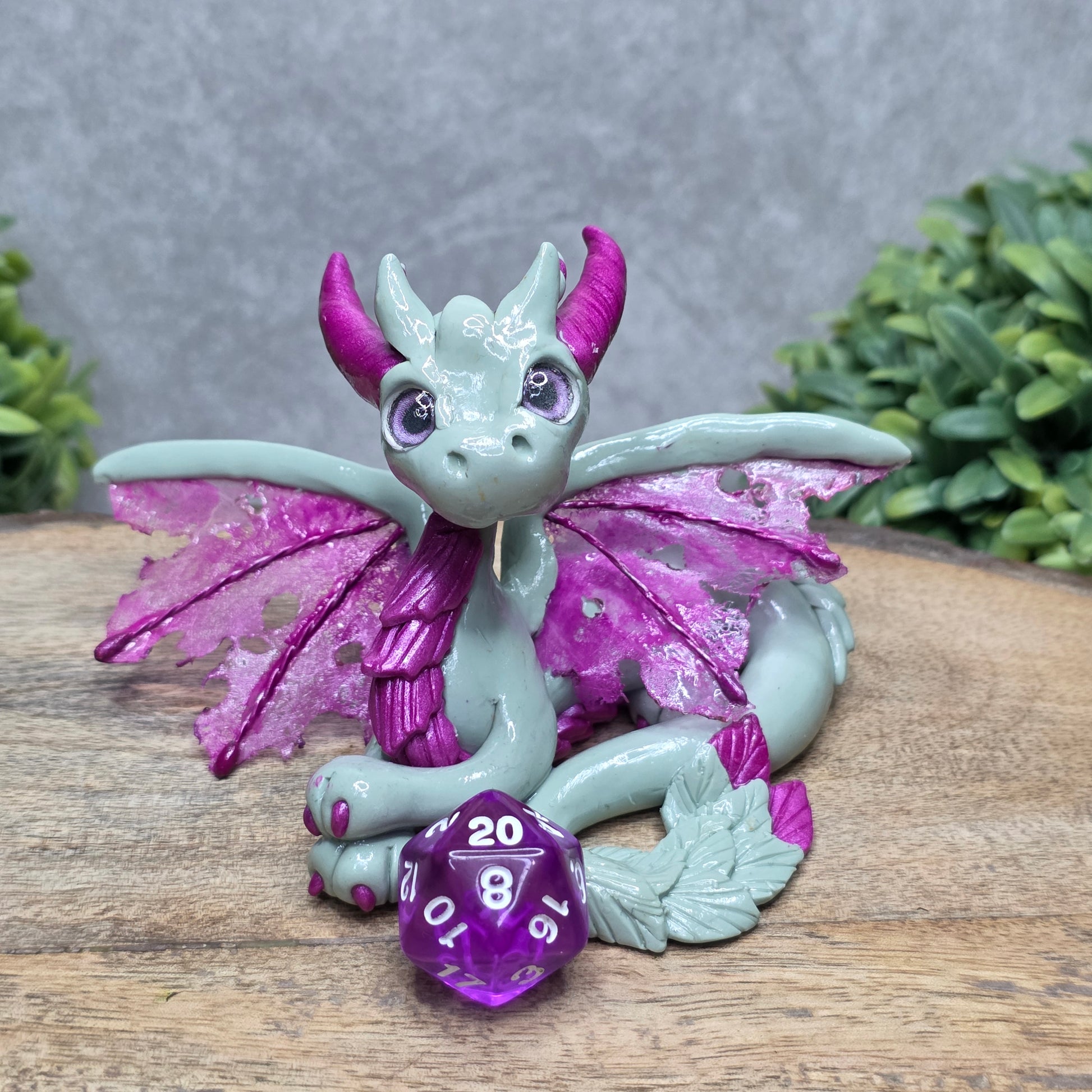 A clay dragon with a D20 dice against a wood surface with greenery.
