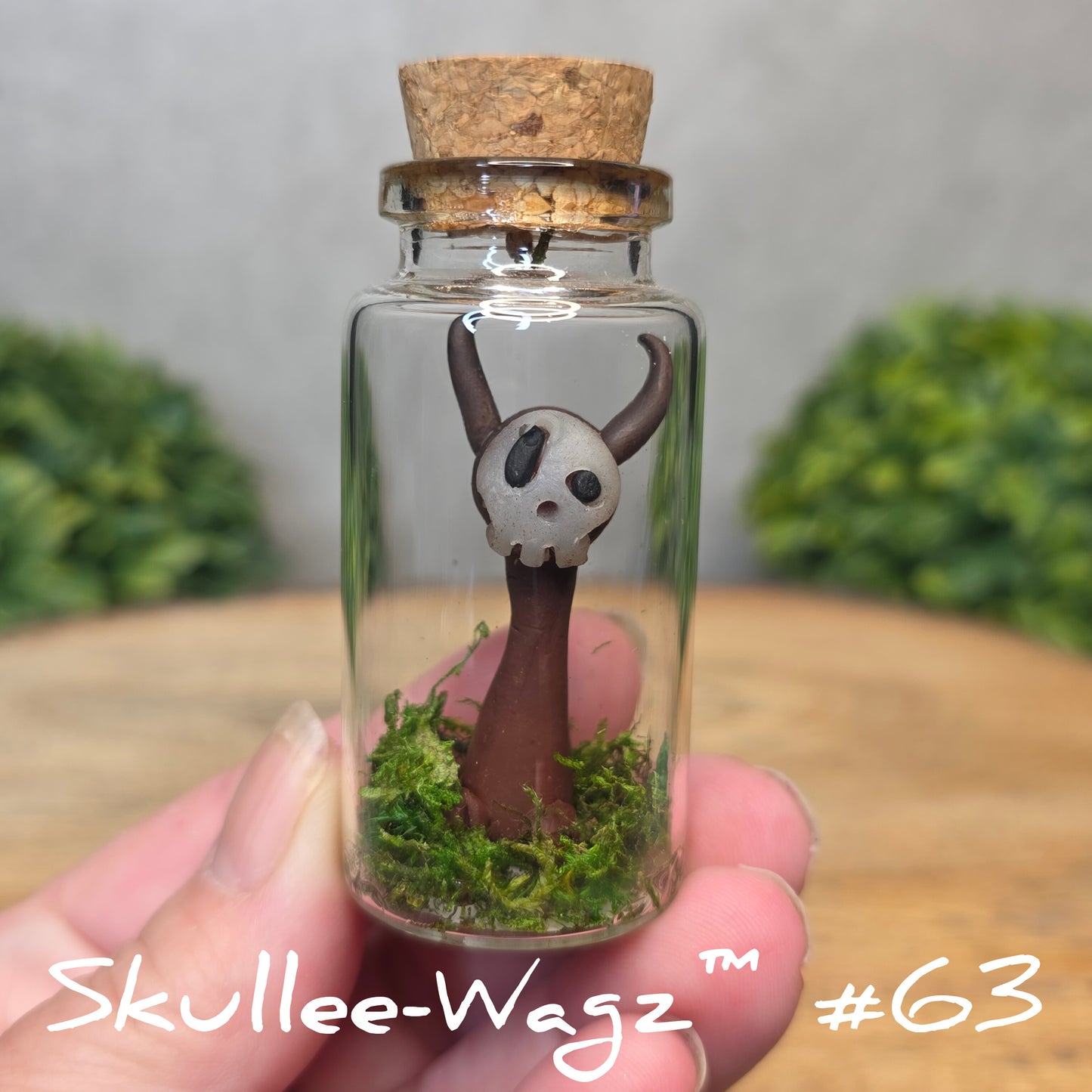 Hand holding a skull faced creature in a jar against a wood background with greenery