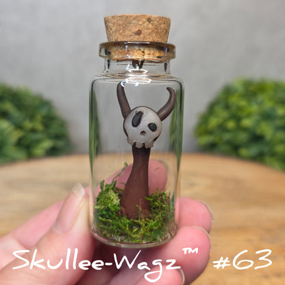 Hand holding a skull faced creature in a jar against a wood background with greenery